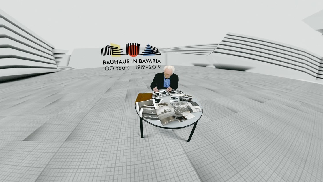 Alexander Cvijanovic sitting in a white architectural room at a table with historical photographs, the Bauhaus in Bavaria 100 Years logo above