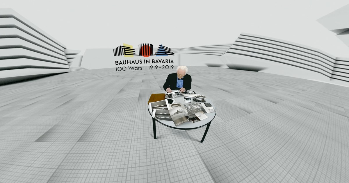 Alexander Cvijanovic sitzt in einem weissen Architekturraum an einem Tisch mit historischen Fotografien, darueber das Logo Bauhaus in Bavaria 100 Years