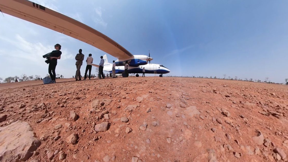 Frachtflugzeug auf einer roten Erdpiste in den Nuba-Bergen im Sudan, Menschen mit Gepaeck davor, 360-Grad-Filmstill