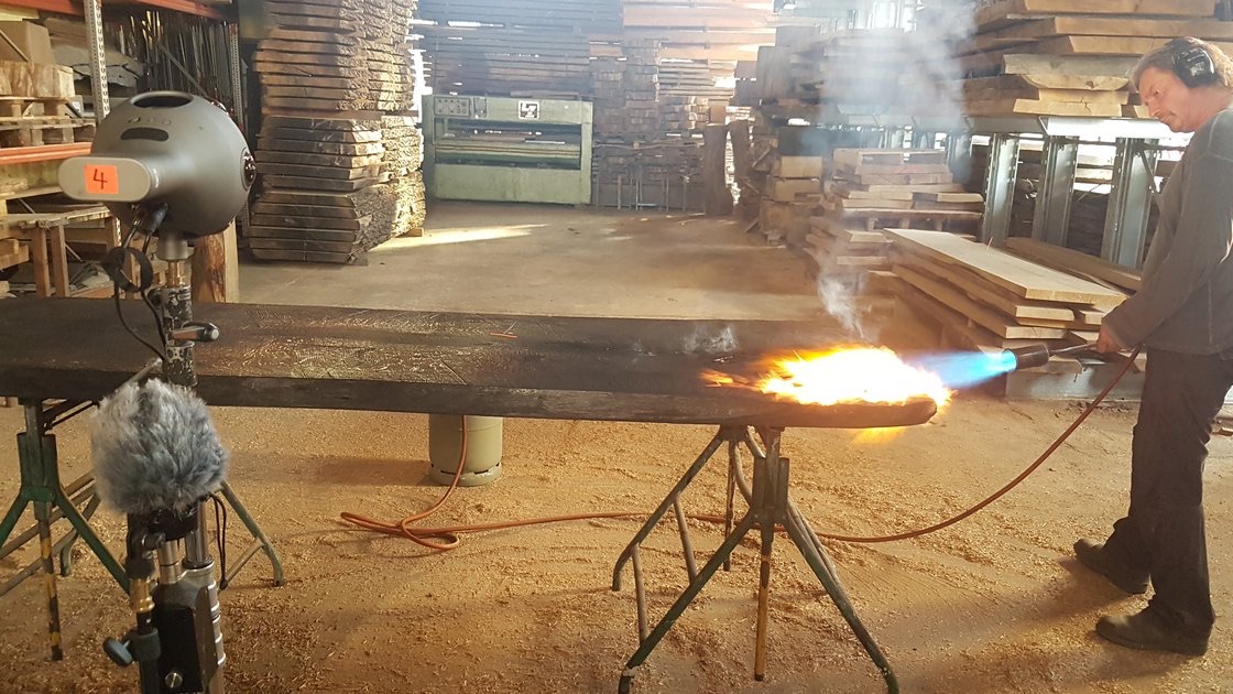 Handwerker behandelt ein Holzbrett mit einer Flamme, im Vordergrund eine 360-Grad-Kamera und ein Mikrofon