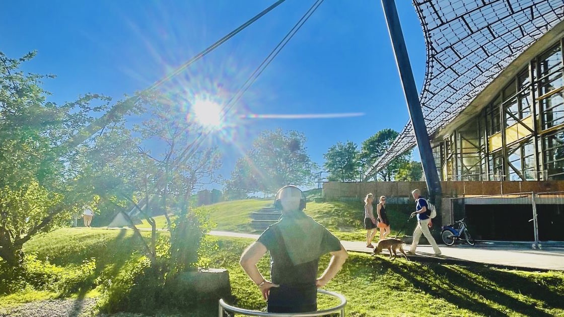 Visitor with a VR headset and headphones on a round standing platform in front of the tent roof of the Olympiapark