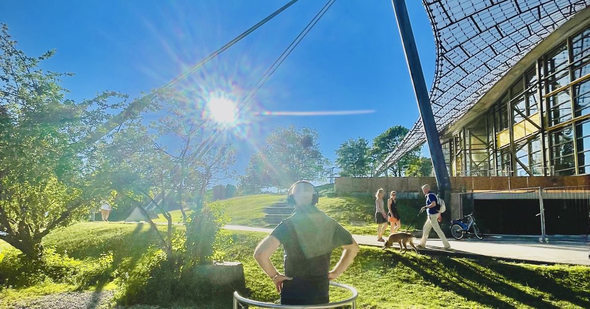 Besucherin mit VR-Brille und Kopfhoerern auf einer runden Standplattform vor dem Zeltdach des Olympiaparks