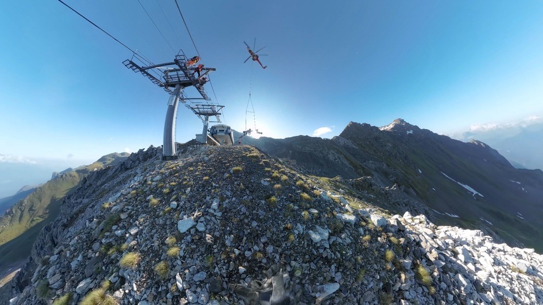360-degree shot on a summit ridge: a helicopter flying a roller battery to a ropeway pylon, the curved horizon showing the 360-degree perspective