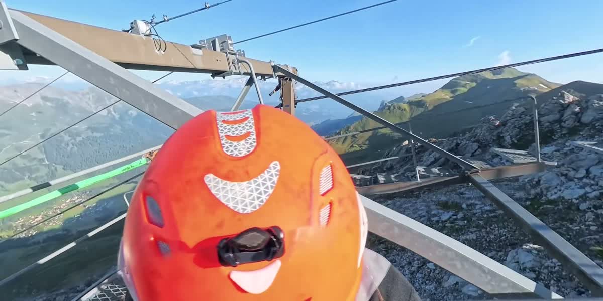 Ropeway mechatronics technician with an orange helmet on the steel structure of a pylon, mountain panorama behind, film still from the 360-degree video